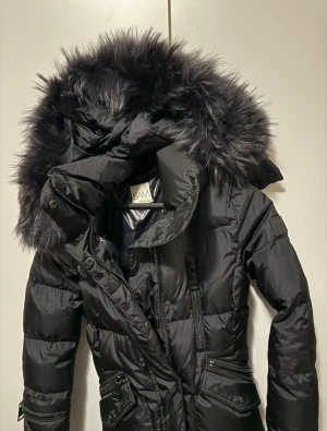 Svart pufferjacka med päls från SAM - Svart pufferjacka från SAM med stor fluffig huva och pälsdetalj. Jackan har flera dragkedjor, fickor med knappar och quiltad design. Perfekt för kalla dagar med sin tjocka och isolerande polyester. Snygg och funktionell look. Märke: SAM • Storlek: XS • Färg: svart • Skick: mycket fint skick • Pris: 1250 kr