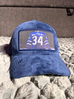 Blå keps från Wristwear med Matthews-tryck - Säljer en blå keps från Wristwear med ett stort tryck på framsidan som visar ryggen på en hockeytröja med namnet Matthews och nummer 34. Kepsen har mjuk, sammetsliknande yta och mesh på insidan för ventilation. Perfekt för dig som gillar sportig stil.