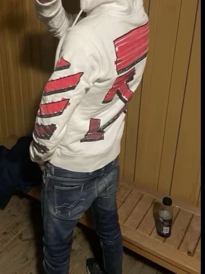 Vit Off-White hoodie  - Säljer en vit hoodie från Off-White med ikoniska grafiska röda och svarta tryck på ärmar och rygg. Tröjan har huva med snörning och en stor magficka. Perfekt statement-plagg för dig som gillar streetwear och unika prints.                                 !!!!!Har äkthetsbevis från vestaiere med en stämpel från fabriken med legit check !!!!