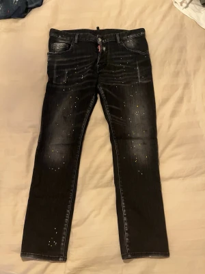 Svarta Dsquared2 ICON jeans med stänk - Säljer ett par svarta Dsquared2 ICONIBRAHIMOVIC jeans i storlek 48 med coola vita och gula färgstänk på både fram- och baksida. Jeansen har klassisk femficksdesign, raka ben och tydlig Dsquared2-label bak. Materialet är jeans och passformen är normal.