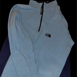 Ljusblå fleece från The North Face - Ljusblå fleece-tröja från The North Face med svart logga på bröstet och mörkblå paneler i sidorna. Tröjan har half zip och hög krage, perfekt för kyliga dagar. Tillverkad i mjuk Polartec-fleece och har långa ärmar.