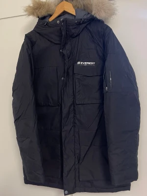 Svart parkajacka från Everest med fuskpäls - Svart parkajacka från Everest med stor huva och fuskpäls. Jackan har flera praktiska fickor, dragkedja och knappar framtill. Materialet är polyester och jackan är fodrad för extra värme. Perfekt för kalla vinterdagar och har en snygg, klassisk look.