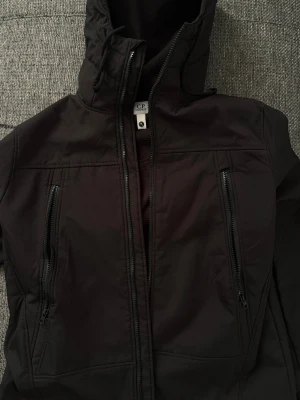 Svart vindjacka från C.P. Company - Svart vindjacka från C.P. Company med ikoniska goggles i huvan. Jackan har två vertikala fickor med dragkedja, hög krage med snörning och mjukt fleecefoder på insidan. Det finns inga skador på jackan topp skick passar bra på storlek M också. Pris kan diskutera. 