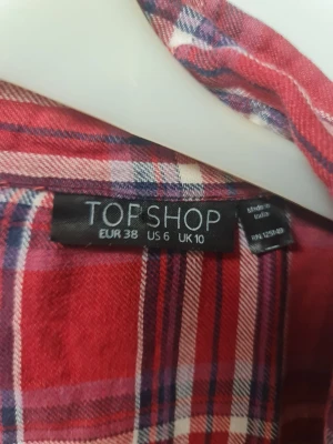 Röd rutig skjorta från Topshop - Säljer en röd rutig skjorta från Topshop i storlek 38. Skjortan har klassisk krage, långa ärmar och knappar framtill. Materialet är mjuk bomull och mönstret är rutor i rött, vitt och blått. Perfekt för dig som gillar avslappnad och cool stil.