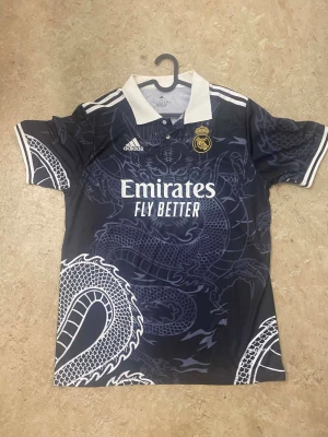 Real Madrid x Adidas draktröja - Säljer en svart Real Madrid x Adidas matchtröja med vit krage och detaljer. Tröjan har ett stort drakmönster i mörkblått och vitt över hela plagget, samt klubbmärke och Adidas-logga på bröstet. Perfekt för dig som vill sticka ut på läktaren eller i stan.