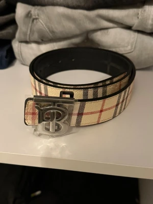 Rutig Burberry bälte med B-spänne - Säljer ett snyggt Burberry bälte med klassiskt rutig design i beige, svart och röd. Bältet har ett metallspänne med ett stort B framtill. Materialet är skinn och mönstret är typiskt Burberry. Perfekt för att lyfta din outfit. OBS STORLEK ÄR 105CM