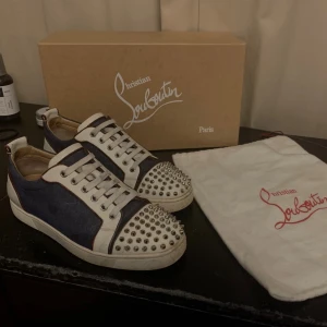 Christian Louboutin sneakers med nitar - Säljer ett par Christian Louboutin sneakers i mörkblått och vitt med coola silvriga nitar på tån. Skorna har klassisk röd sula, snörning och detaljer i skinn. Kommer med originalkartong och dustbag. Perfekt för dig som vill sticka ut med lyxiga och edgy sneakers. Saknar bara en nit annars mycket bra