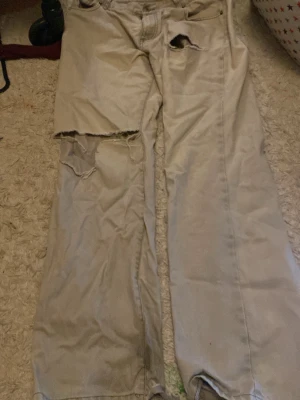 Beige lowrise  jeans med hål  - Säljer ett par beige lowrise jeans med klassisk femficksdesign och raka ben. Jeansen har slitna detaljer och stora hål på ena benet och vid fickan, vilket ger en rå och edgy vibe. Materialet är robust bomull och passformen är straight.