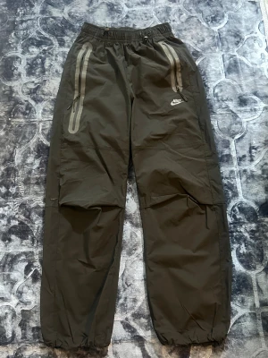 Nike svarta byxor med dragkedja XS - Nike byxor i svart med elastisk midja och dragsko, två framfickor med dragkedja och reflekterande detaljer. Materialet är syntet och byxorna har en sportig look med Nike-logga på framsidan. Passar dig som gillar streetwear och funktionella plagg. Byxorna är helt nya och absolut oanvända med prislapp. Pris går att diskutera vid snabb affär! 