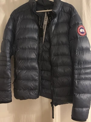 Mörkblå dunjacka från Canada Goose - Säljer en mörkblå dunjacka från Canada Goose med klassisk patch på ärmen. Jackan har quiltad design, dragkedja framtill och långärmade ärmar. Perfekt för kalla dagar och har en snygg, sportig look med diskret glans i materialet.