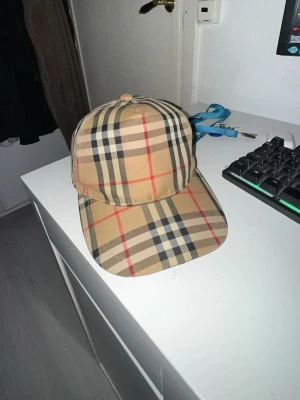 Rutig Burberry keps i bomull - Säljer en klassisk Burberry keps med det ikoniska rutmönstret i beige, svart, vitt och rött. Kepsen har justerbar snapback och Burberry London England-logga bak. Priset kan diskuteras(;