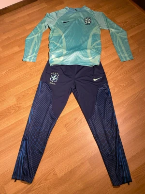Nike Brasil träningsset blå/grön - Nike Brasil träningsset med långärmad tröja i ljusgrönt med grafiskt mönster och halv dragkedja, samt marinblå träningsbyxor med ljusgröna detaljer och Brasil-logga. Perfekt för fotbollsträning och sportiga dagar.