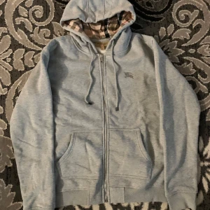 Äkta Grå hoodie från Burberry - Säljer en stilren grå hoodie från Burberry med dragkedja och klassiskt rutigt mönster i huvan. Tröjan har en broderad logga på bröstet och praktiska fickor framtill. Perfekt för en avslappnad och trendig look.