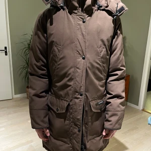 Brun parkajacka med huva - Canada Goose - Säljer en brun parkajacka från Canada Goose med stor huva och praktiska fickor framtill. Jackan har dragkedja och knappar, samt justerbar midja. Är av äldre modell men är utan slitage, från ett djur- och rökfritt hem. Perfekt för kalla dagar och har en clean, stilren look. Dunfyllning. Enligt etiketten är det en L men upplevs som M. Kanske är ryggen med sitt indrag mer som dam-modell men skulle själv beskriva den som en unisex, klassisk modell.