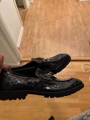 Loafers - Snygga svarta loafers från Jemson med glansig finish och geometriskt mönster. Skorna har guldfärgade metallspännen framtill och en robust, räfflad sula för extra grepp. Perfekt för dig som gillar stilrena detaljer och vill sticka ut.