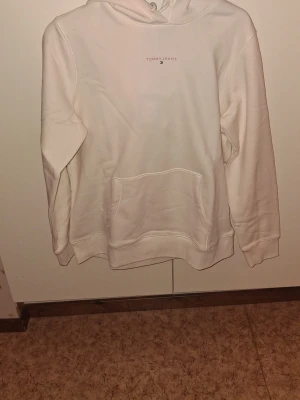 Tommy jeans hoddie  - Tommy Jeans hoodie i helt nytt skick. Hoodien är aldrig använd och helt fri från fläckar, slitage eller skador. Den är vit med snygg rosa text, vilket ger ett fräscht och modernt intryck. Klassisk Tommy Jeans-design med hög kvalitet och bekväm passform. Storlek L, passar perfekt till både vardag och fritid. Ett toppenplagg för dig som vill ha en helt ny hoodie i original.