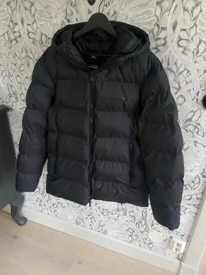 Svart pufferjacka från J.Lindeberg - Säljer en svart pufferjacka från J.Lindeberg med huva och diskret logga på bröstet och ärmen. Jackan har dragkedja framtill och två sidofickor med dragkedja. Jackan är i ny skick och har inga hål eller fläckar!