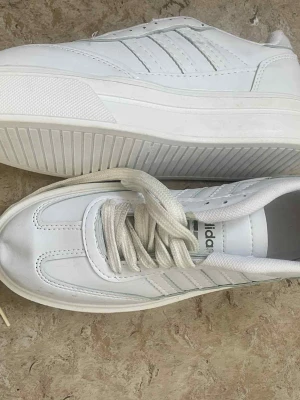 Vita adidas POD S3.1 sneakers - Säljer ett par fräscha vita adidas POD S3.1 sneakers med klassiska tre ränder på sidan och adidas-logga på plösen. Skorna har snörning och en platt sula. Materialet är syntet och skinn, vilket ger en clean och modern look.