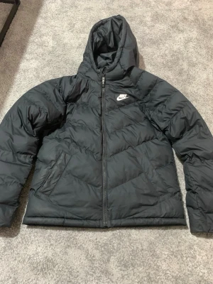 Svart pufferjacka från Nike XL - Svart pufferjacka från Nike i storlek XL, med huva och dragkedja framtill. Jackan har ett stilrent Nike-logga på bröstet och två sidofickor. Materialet är polyester och jackan är perfekt för kalla dagar. Passformen är normal och ärmarna är långärmade.