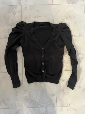 Svart kofta från Vero Moda - Säljer en svart kofta från Vero Moda med puffade axlar och v-ringad hals. Koftan har långa ärmar och knappar framtill. Materialet är mjukt och känns som bomull eller syntet. Perfekt att slänga på sig när det är lite kyligt ute.