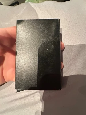 Svart plånbok i metall - Säljer en stilren svart plånbok i metall med slät yta och rektangulär form. Insidan har röda detaljer och fack för kort. Perfekt för dig som vill ha en enkel och modern plånbok som sticker ut lite extra.