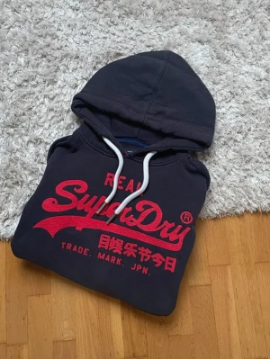 Svart SuperDry hoodie - Hej! Säljer en svart Superdry hoodie i storleken L. Hoodien är har tre små slitningar på baksidan av höger ärm (se bild). Utöver detta är hoodien i bra skick.