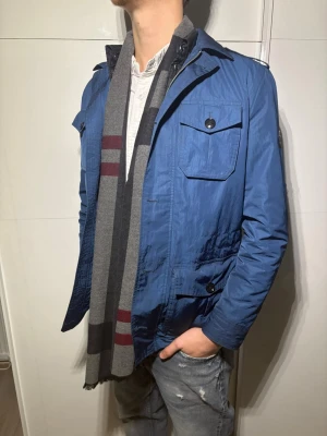 Blå vindjacka från Lindberg - Snygg blå vindjacka från Lindberg med klassisk krage, flera fickor med knappar och en cool patch på ärmen. Jackan har dragkedja och knappar framtill, i jackan så finns det även en väst som tillhör jackan som du kan ta på och av. Skick 10/10🤩