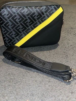 Fendi axelväska med gul detalj - Säljer en snygg axelväska från Fendi med klassiskt Fendi-mönster i svart och samt en diagonal gul skinnremsa. Väskan har en bred, svart axelrem med Fendi-logga och silverfärgade metalldetaljer. Perfekt för dig som vill sticka ut med stil.