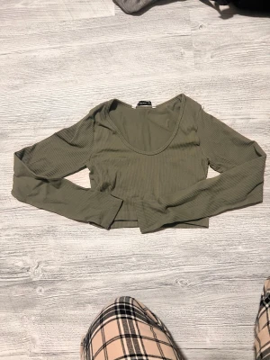 Olivgrön ribbad croppad topp - Säljer en olivgrön croppad topp med lång ärm och ribbad struktur. Toppen har en rundad, lite djupare halsringning och är gjord i mjuk bomullsmix. Perfekt att styla med höga jeans eller kjol.