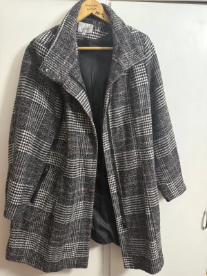 Rutig kappa från Isolde, storlek 44 - Snygg rutig kappa från Isolde i svart, vit och diskret rosa. Jackan har hög krage, dragkedja och fickor med zip. Materialet är mestadels polyester med inslag av akryl och blandade fibrer, fodret är helt i polyester. Perfekt för dig som gillar klassisk stil med en twist.