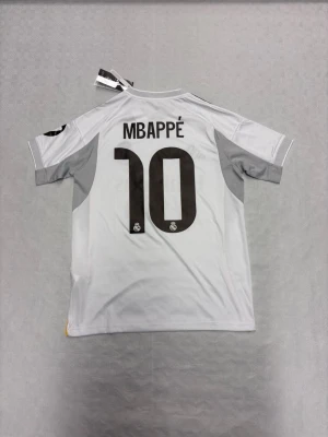 Real Madrid Mbappé Tröja 25/26 -  En officiell Real Madrid matchtröja med Mbappé och nummer 10 på ryggen. Tröjan är vit med svarta och gula detaljer, broderat klubbmärke och Adidas-logga. Materialet är lätt top kvalité  med Aeroready-teknologi och svarta ränder på axlarna.