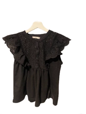 Svart blus med spets och volang från MNG - Supersöt svart blus från MNG med broderade blommor och spetsdetaljer på axlarna. Blusen har volang vid axlarna, knappar framtill och är ärmlös. Perfekt att styla med jeans eller kjol för en trendig look.