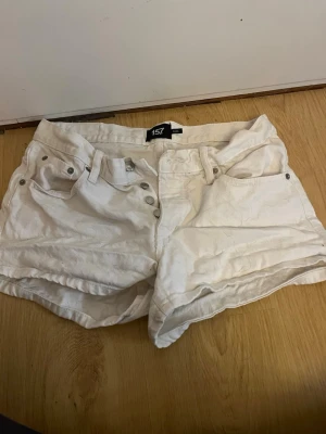 Vita shorts från 157 med broderi - Säljer ett par vita shorts från 157 med snygga broderade detaljer på bakfickorna. De har låg midja, knappgylf och klassisk femficksdesign. Materialet är bomull och de har bälteshällor för extra stylingmöjligheter.
