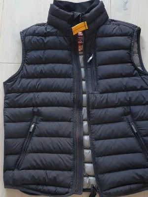 Svart dunväst från Parajumpers - Säljer en svart dunväst från Parajumpers med quiltad design och hög krage. Västen har dragkedja framtill, två snedställda fickor med dragkedja och en gul detalj vid halsen. Insidan är ljusgrå och materialet är polyester med dunfyllning.