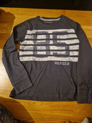 Mörkblå långärmad topp Tommy Hilfiger - Säljer en mörkblå långärmad topp från Tommy Hilfiger i mjuk bomull. Framsidan har ett stort tryck med vita och blå ränder samt Hilfiger-logga. Klassisk rund hals och diskret logga på ärmslutet. Perfekt för dig som gillar stilrena och sportiga plagg.