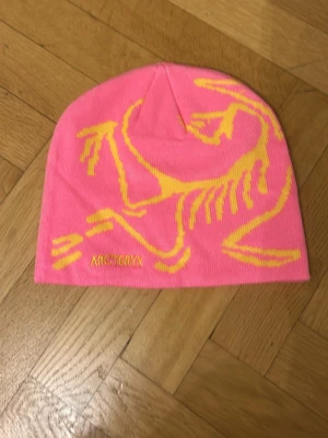 Rosa Arc’teryx mössa - Bilderna visar en rosa mössa från Arc'teryx med gul logga och motiv, inte en väst. Kan ej ge västbeskrivning eller taxonomier.
