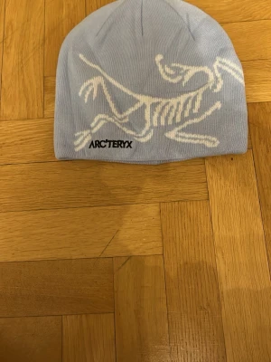 Ljusblå mössa från Arc'teryx - Snygg ljusblå mössa från Arc'teryx med vit fossil-logga på framsidan. Mössan är stickad och har en enkel, rundad form som sitter skönt på huvudet. Perfekt för kalla dagar och outdoor-aktiviteter.