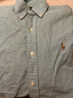 Ljusblå skjorta från Polo Ralph Lauren - Snygg ljusblå skjorta från Polo Ralph Lauren med klassisk krage och knappar framtill. Ikonisk broderad logga med polospelare på bröstet. Skjortan är tillverkad i mjuk bomull och har en stilren, tidlös look som passar perfekt till jeans eller chinos.