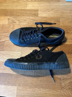 Svarta Fred Perry sneakers - Säljer ett par svarta sneakers från Fred Perry med klassisk logga på sidan. Skorna har snörning, rund tå och en rejäl gummisula med mönster undertill. Ovandelen är i textil och detaljer i gummi vid tå och häl. .