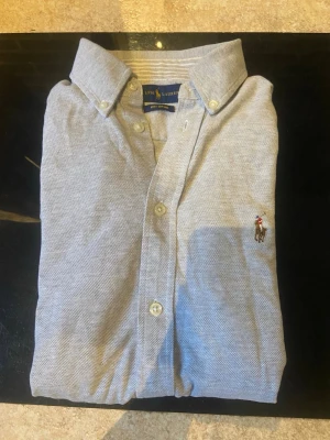 Ljusgrå oxfordskjorta från Ralph Lauren - Snygg ljusgrå oxfordskjorta från Ralph Lauren med klassisk krage och knappar framtill. Skjortan har det ikoniska broderade Polo-logot på bröstet och är tillverkad i mjuk bomull. Perfekt för dig som gillar stilrena och tidlösa plagg.