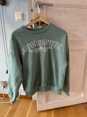 Grön sweatshirt Dufourspitze M - Mjuk grön sweatshirt från Dufourspitze med broderad vit text framtill. Tröjan har rund hals, ribbade muddar för extra komfort. Sliten invändigt vilket syns på bilderna. Perfekt för chill dagar och enkel att matcha med jeans eller mjukisbyxor.