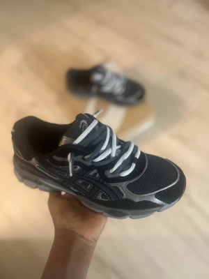 Svarta och grå Asics sneakers - Säljer ett par snygga Asics sneakers i svart och grått med dubbel snören. Skorna har mesh- och syntetpaneler, rund tå och platt sula. Perfekta för dig som gillar sportig stil och vill ha bekväma skor till vardagen. Box ingår. Man kan också ta bort en av snören om man vill och priset kan diskuteras.
