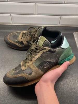Valentino rockrunners  - Säljer ett par Valentino Garavani Rockrunner sneakers med camouflage-mönster i grönt, beige och svart. Skorna har snörning, platt sula och detaljer i mocka och skinn. Baktill finns mintgröna och gröna partier med nitar. Perfekt för dig som gillar streetstyle och designer.