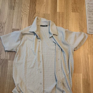 Beige kortärmad skjorta från 157 - Säljer en beige kortärmad skjorta från 157 med krage och knappar framtill. Skjortan har en lätt ribbad struktur i tyget och är i en luftig modell, perfekt för varmare dagar. Enkel och stilren design som är lätt att matcha.
