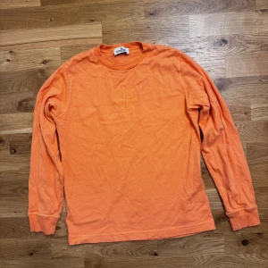 Orange långärmad tröja Stone Island - Orange långärmad tröja från Stone Island Junior med rund hals och broderad logga på bröstet. Tröjan är i mjuk bomull och har ribbade muddar vid ärmslut och hals. Perfekt för dig som gillar streetwear och vill sticka ut med färg.