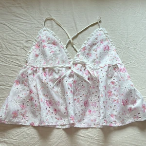 Vit blommig linnetopp - Supersöt vit linnetopp med broderade blommor i rosa och små hålbroderier. Toppen har smala justerbara axelband som korsas i ryggen, volangkant vid bysten och knyt framtill. Materialet är 100% polyester och toppen är fodrad. Perfekt för sommarens alla tillfällen! 💕🤍