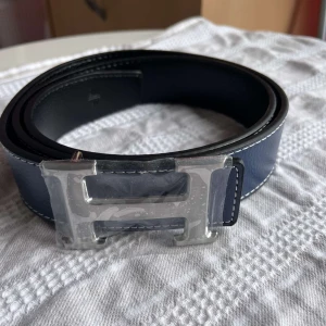 H-bälte  - Snyggt bälte i blått och svart skinn med ikoniskt,H-spänne i silverfärgad metall. Bältet har stilrena, kontrastsömmar och klassisk design som lyfter, vilken outfit som helst. Strl 110cm