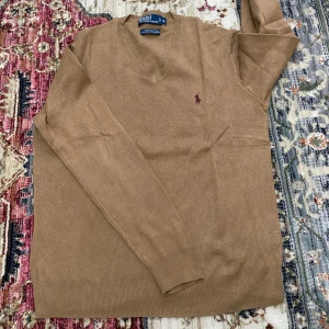 Tröja från Polo Ralph Lauren - Snygg beige v-ringad tröja från Polo Ralph Lauren i 100% pima bomull. Tröjan har lång ärm och den klassiska broderade loggan i vinrött på bröstet. 