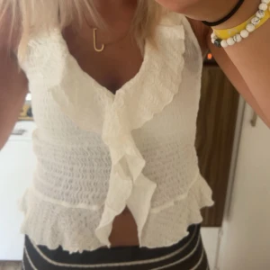 Vit volangblus med omlottkänsla - Supersöt vit blus med volangkant och omlottkänsla framtill. Smockad struktur ger en snygg passform och den är ärmlös, perfekt för varma dagar. Volangerna ger en romantisk vibe och toppen är lätt att matcha med både jeans och kjol. Knappast använd