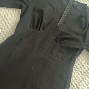 Svart långärmad klänning från SHEIN - Svart klänning från SHEIN med lång ärm och markerad midja. Klänningen har v-ringad omlott-look upptill, diskreta rynkdetaljer vid bysten och ärmslut samt dragkedja i ryggen. Materialet är mjukt och har en lätt glansig finish.